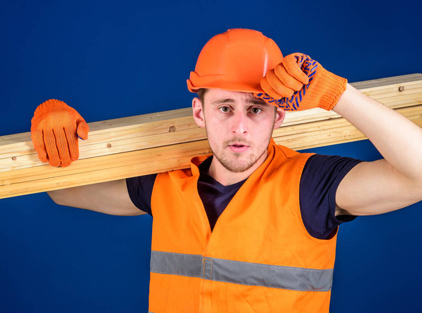 Man in protective gloves holds visor of helmet, corrects hard hat on head, blue background. Carpenter, woodworker, labourer, builder carries wooden beam on shoulder. Woodworker concept - Фото, зображення