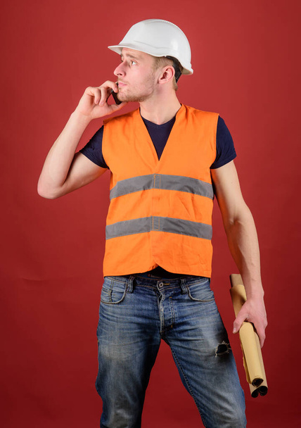 Architect calling engineer to fix plan. Man, foreman in helmet speaking on phone, red background. Engineer, architect, on busy face speaks on smartphone while holds blueprints. Communication concept. - Photo, Image