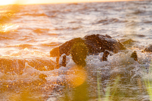 Ondas quebram em uma costa de quebra-mar pedregosa e formam um grande spray com cores de pôr do sol e brilho da lente
 - Foto, Imagem
