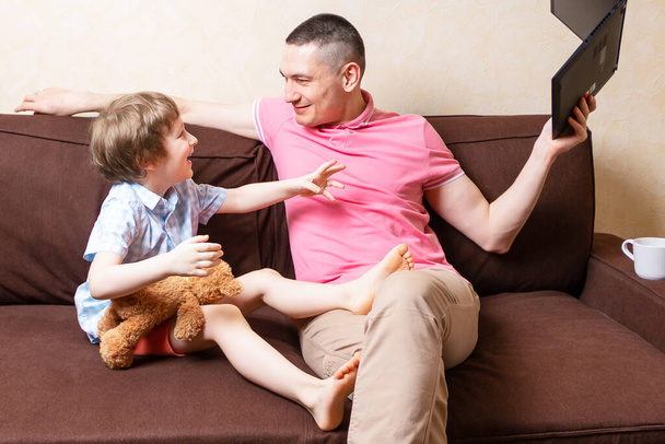 Hauska nuori isä istua tietokoneen kannettavan pelleillä söpö lapsi poika pitää hauskaa yhdessä, hymyilevä isä ja pieni poika lapsi nauttia pysyä kotona levätä sohvalla vempaimia. Online koulutus ja työ
 - Valokuva, kuva