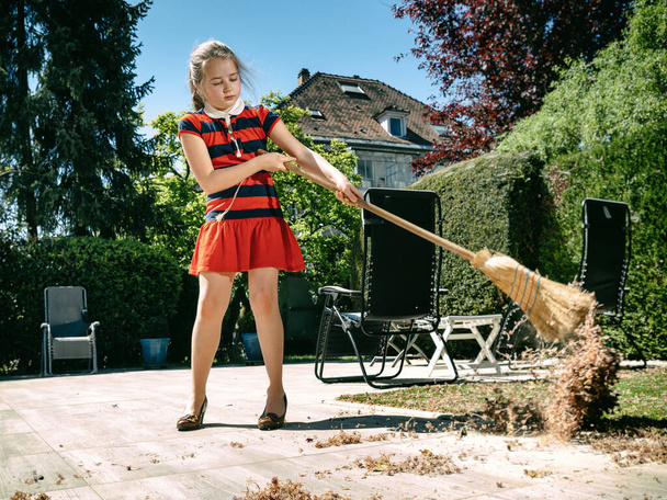 A schoolgirl poses with a broom and in her mothers shoes in the yard.  Sweeping in the garden. Sunny weather. Joyful childhood. - Foto, Imagen