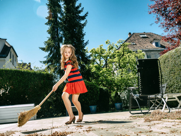 A schoolgirl poses with a broom and in her mothers shoes in the yard.  Sweeping in the garden. Sunny weather. Joyful childhood. - 写真・画像
