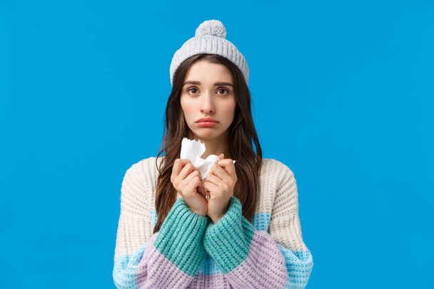 Upset young woman feelings unwell, have fever, sick on winter holidays, wearing hat and sweater, holding napkin sneeze and have runny nose, looking sad camera, caught cold or flu, blue background - Фото, изображение