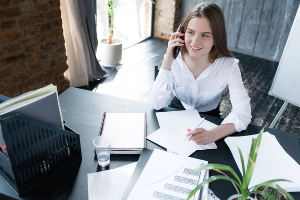 American woman talking on a smartphone and smiling. The secretary works in a modern office and contacts clients - Photo, image