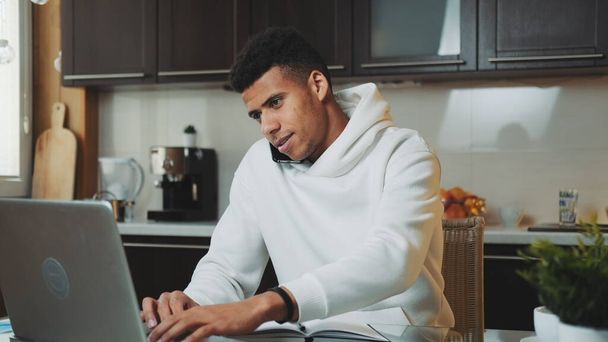 Vegyes fajú férfi beszél okostelefonon, és dolgozik a számítógép az otthoni konyhában - Fotó, kép