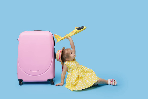 Cute child girl in summer dress with a pink suitcase and toys airplane is dreams of traveling to tropical countries after quarantine. Blue studio background.  - 写真・画像