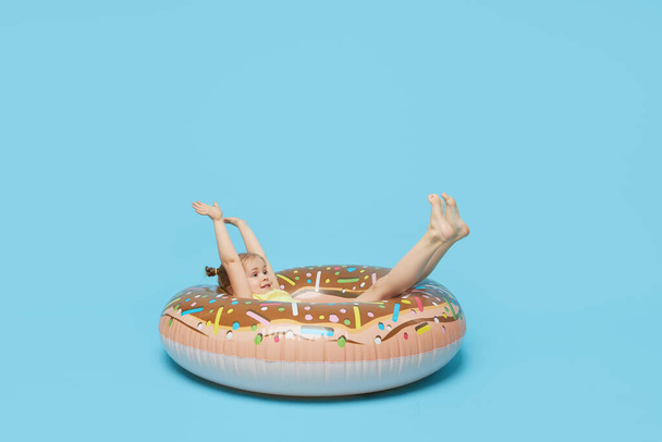 Failed summer vacation. Happy surprised child model girl resting on a donut-shaped rubber ring. Sea vacation concept on a colored blue background.  - Fotoğraf, Görsel