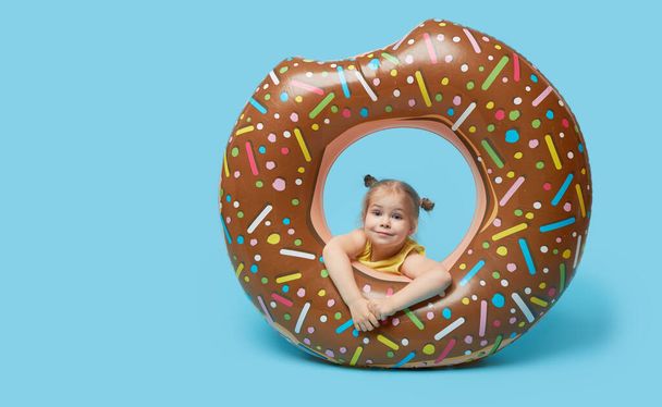 Failed summer vacation. Happy surprised child model girl resting on a donut-shaped rubber ring. Sea vacation concept on a colored blue background.  - Foto, Bild