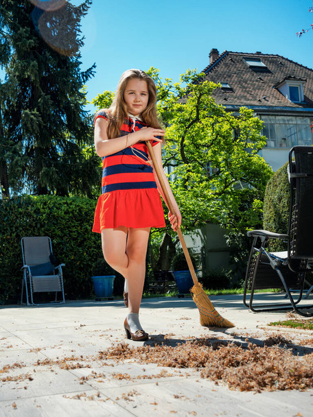 A schoolgirl poses with a broom and in her mothers shoes in the yard.  Sweeping in the garden. Sunny weather. Joyful childhood. - Photo, image