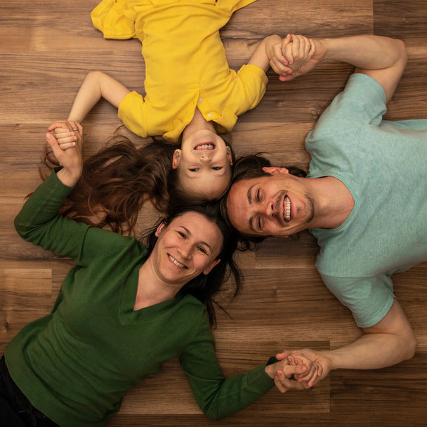 Widok z góry młodych rodziców i dziewczynki patrzącej w kamerę i uśmiechniętej leżącej na podłodze w domu. - Zdjęcie, obraz