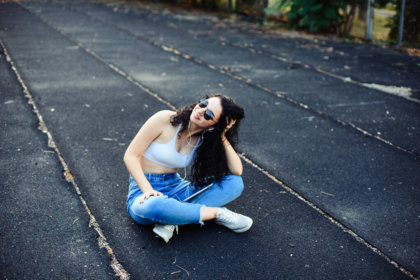 A young brunette with glasses with long curly hair sits on the ground in the street in a white top and jeans with a tablet in her hands and headphones in her ears. Life style. - Foto, Imagem