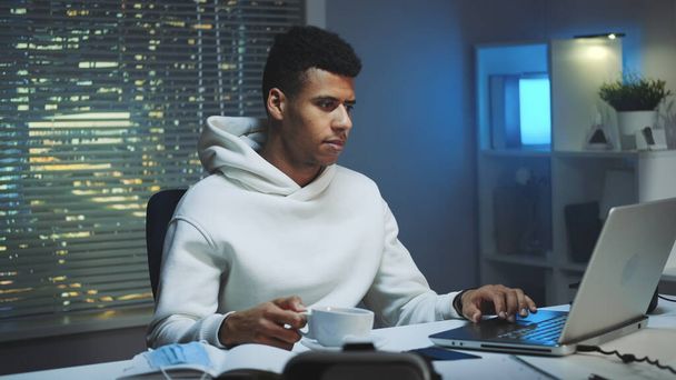 Handsome multiracial man drinking a cup of coffee and working on computer at night - Photo, Image