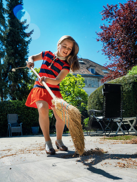 A schoolgirl poses with a broom and in her mothers shoes in the yard.  Sweeping in the garden. Sunny weather. Joyful childhood. - Φωτογραφία, εικόνα