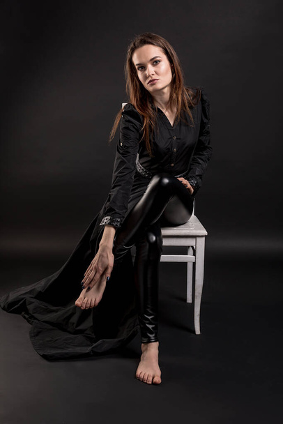 A makeup brunette girl in dark clothes is sitting on a chair. Studio portrait dark background. Vertical orientation. High quality photo. - 写真・画像