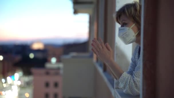 Coronavirus pandemia maailmassa, ihmiset jäävät kotiin, karanteeni käsite. Nainen suojelevassa naamiossa katselee ikkunasta ja taputtaa käsiään tukemalla kansallista flash-väkijoukkoa Italiassa, taputtaen
 - Materiaali, video
