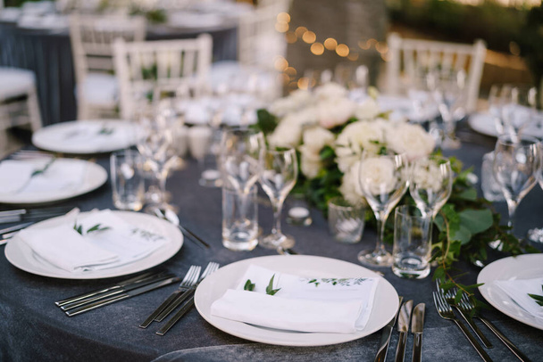 Wedding dinner table reception. White round plates on a round table with gray tablecloth, white Chiavari chairs with white pillows. A floral arrangement in the center of the table. - Photo, Image