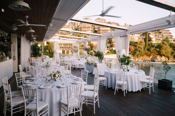 Svatební hostina u stolu. Spousta kulatých stolů s bílými ubrusy a bílými Chiavari židlemi. Restaurace bez střechy s výhledem na moderní vilový komplex v Černé Hoře. Svatba na moři, při západu slunce - Fotografie, Obrázek