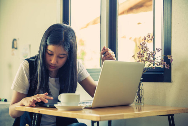 Fiatal nő ül a kávézóban az asztalnál, abbahagyja a munkát a laptopjával, és elveszi az okos telefonját. A lány vezeték nélküli internetet használ. - Fotó, kép