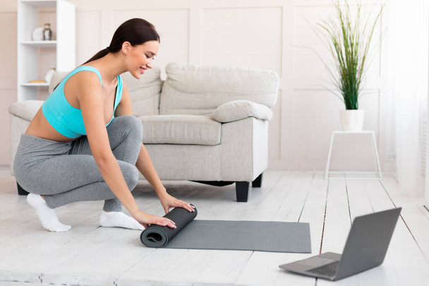 Женщина после успешной тренировки на ноутбуке скручивая коврик в помещении
 - Фото, изображение