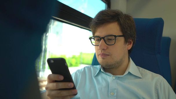 Handsome young businessman wearing a blue shirt and glasses is riding a train dialing a number and talking on his phone. Sunny summer day. - Fotó, kép