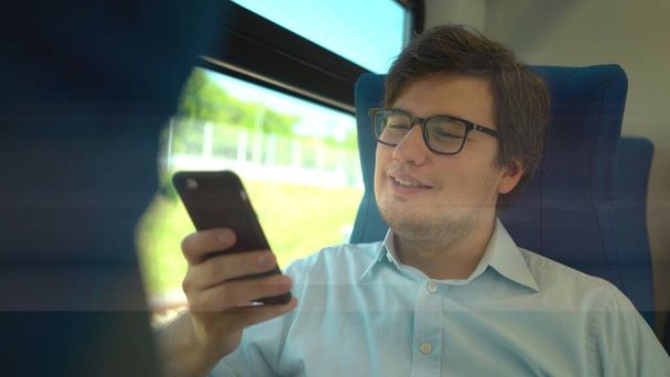 Handsome young businessman wearing a blue shirt and glasses is riding a train dialing a number and talking on his phone. Sunny summer day. - Zdjęcie, obraz