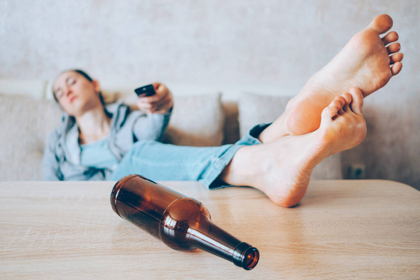 Ένα μεθυσμένο κορίτσι κάθεται σε έναν καναπέ και αλλάζει το κανάλι της τηλεόρασης με ένα μπουκάλι στο επίκεντρο. Κλείσε.. - Φωτογραφία, εικόνα