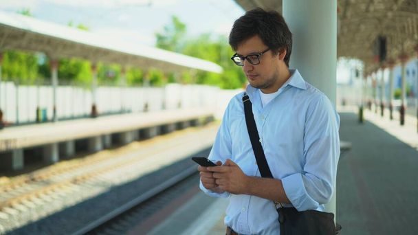 青いシャツと眼鏡を身に着けている若いビジネスマンは、彼の列車を待っているプラットフォームに立っている間、テキストメッセージです。夏の晴れた日.  - 写真・画像