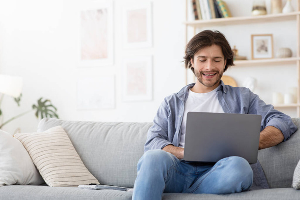 Iloinen kaveri istuu sohvalla kotona, kannettavan tietokoneen avulla
 - Valokuva, kuva