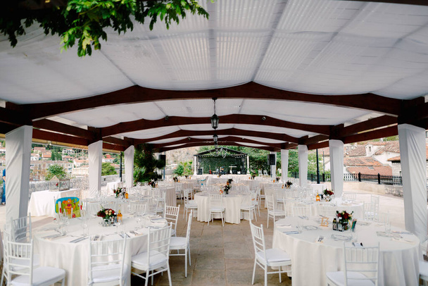 Bruiloft diner tafel receptie. Ronde tafels met witte tafelkleden en witte Chiavary stoelen onder een grote witte tent, tegen de achtergrond van een muzikale groep. - Foto, afbeelding