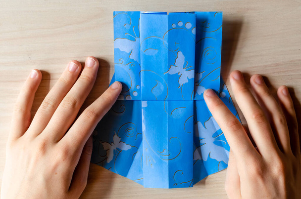 A lány kezei origami kék ruhát készítenek pillangó nyomtatott papírból. Lépésről lépésre, 13. lépés. Anyák napja, születésnap, nyári ajándék, családi szabadidő koncepció. DIY bemutató, lapos, felülnézet. - Fotó, kép
