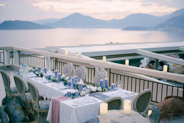 Свадебный ужин с большим количеством густых голубых свечей на фоне закатного неба над горами и морем в Будве, Черногория. Старые мягкие стулья на летней террасе ресторана, на открытом воздухе
. - Фото, изображение
