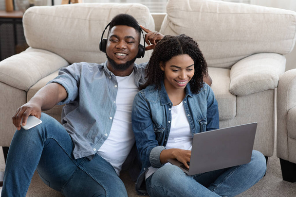 Black Couple Relaxing Together At Home, Listening Music And Using Laptop - 写真・画像
