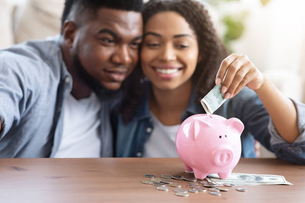 Orçamento Familiar. feliz casal preto colocando dinheiro para Piggybank em casa
 - Foto, Imagem