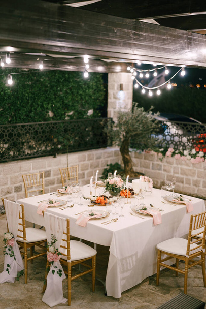 結婚式のディナーテーブルレセプション。白いテーブルクロスとピンクのナプキンを持つ6人のための長方形のテーブル。金色のカイロフキアは布とピンクの花で飾られています。ガーランドはテーブルの上で燃える.  - 写真・画像