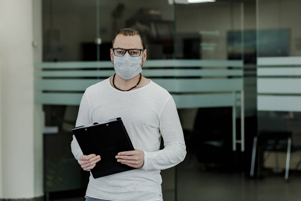 Office worker in glasses wearing a protective mask. Male employee wearing medical facial mask working alone as of social distancing policy. Business and healthcare concept. Copy space. - Foto, imagen