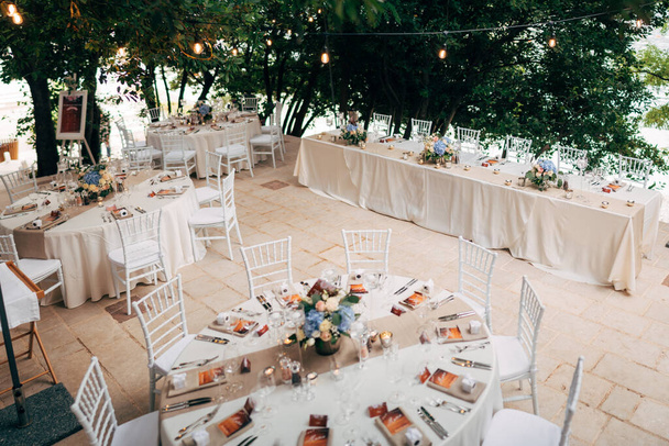 Wedding dinner table reception. Round tables with white tablecloths, brown runners, bouquets flowers, Chiavari chairs, menu sin-enclosed in napkin. Restaurant outside, in woods, water lights garlands - Photo, Image