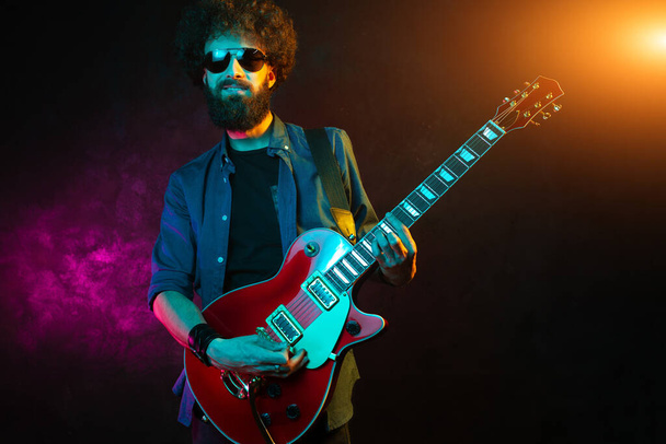 Hipster man with curly hair with red guitar in neon lights. Rock musician is playing electrical guitar. - Foto, Bild