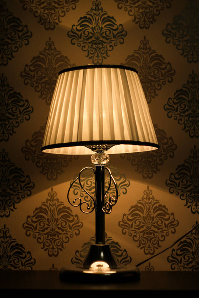 Picture  of a bedside silver lamp, which is standing on a bedside table with a white lampshade at night. It creates a romantic and calm atmosphere. Shades are on the walls with a silver wallpaper - Фото, изображение