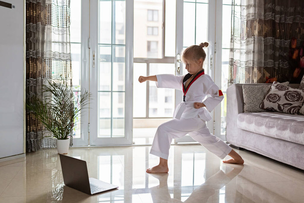 Taekwondo girl in kimono with white belt exercising at home in living room. Online education during coronavirus covid-19 lockdown, self isolation and social distancing concept - Fotó, kép