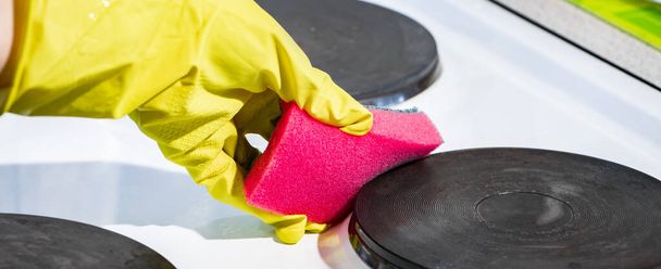 Η διαδικασία πλύσης μιας ηλεκτρικής σόμπας. Ένα κορίτσι με ένα κίτρινο γάντι καθαρίζει τη λευκή επιφάνεια της βρωμιάς. Καθαρισμός εργασιών. Ροζ σφουγγάρι κουζίνας στα χέρια της γυναίκας. Μορφή κλειδιού - Φωτογραφία, εικόνα