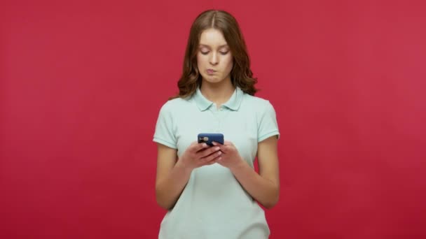 Mobile communication, online chatting. Young brunette woman in polo t-shirt typing message, texting in social network using cell phone, browsing web. indoor studio shot isolated on red background - Metraje, vídeo