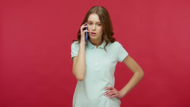 Wow, unbelievable! Amazed young brunette woman in polo t-shirt speaking on mobile phone and expressing surprise at incredible news, chatting with friend. indoor studio shot isolated on red background - Materiał filmowy, wideo