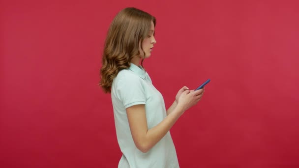 Mobile communication. Side view of brunette woman in polo t-shirt typing message or chatting online using smartphone, expressing surprise at read news. indoor studio shot isolated on red background - Filmagem, Vídeo