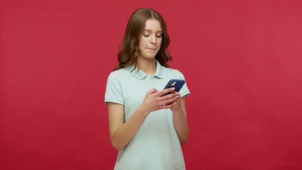 Online mobile communication. Young brunette woman in polo t-shirt typing message, thinking over answer on social network and texting on smartphone. indoor studio shot isolated on red background - Footage, Video