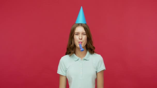 Birthday surprise. Happy young brunette woman in polo t-shirt and funny cone on head blowing party horn and catching gift box, celebrating holidays. indoor studio shot isolated on red background - Video