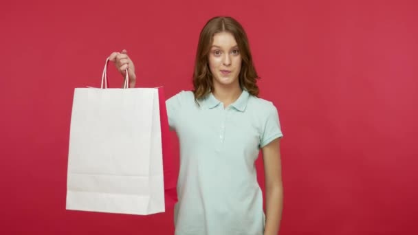 Happy shopper recommending discounts. Young woman in polo t-shirt showing Sale inscription and shopping bags with white empty copy space, satisfied with low cost purchase. indoor studio shot isolated - Video