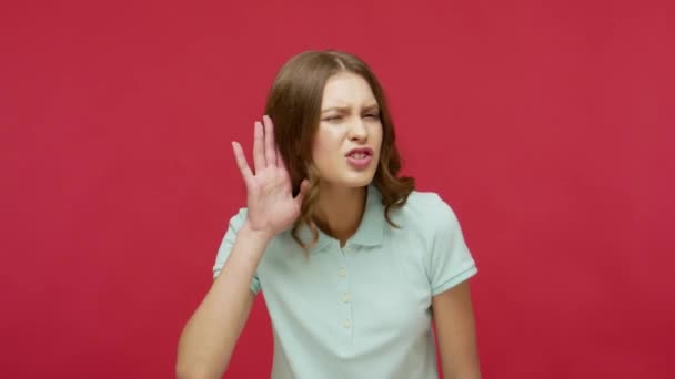 What? I can't hear you! Brunette young woman in polo t-shirt holding hand near ear to listen, suffering from hearing problems, focused on quiet conversation. studio shot isolated on red background - 映像、動画