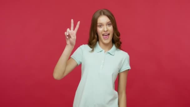 Carefree positive young brunette woman in polo t-shirt having fun and showing victory or peace gesture, double v sign, celebrating winning score, success. indoor studio shot isolated on red background - Filmagem, Vídeo