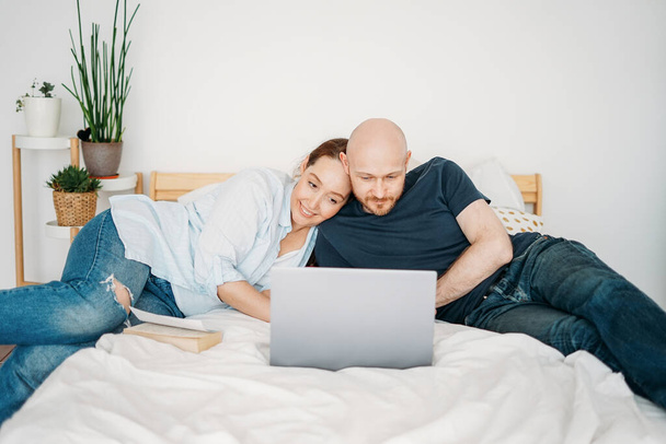 Adult married couple in casual clothing using open laptop lie on the bed at bright interior - Photo, image