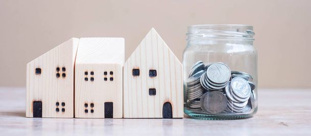 ガラス瓶のコインとテーブルの上の木製の家のモデル。ビジネス、投資、お金の節約、不動産および退職計画の概念 - 写真・画像